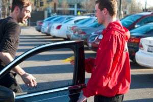 A parking valet attendant wearing a red jacket is handing a yellow piece of paper to a man stepping out of a black vehicle. 